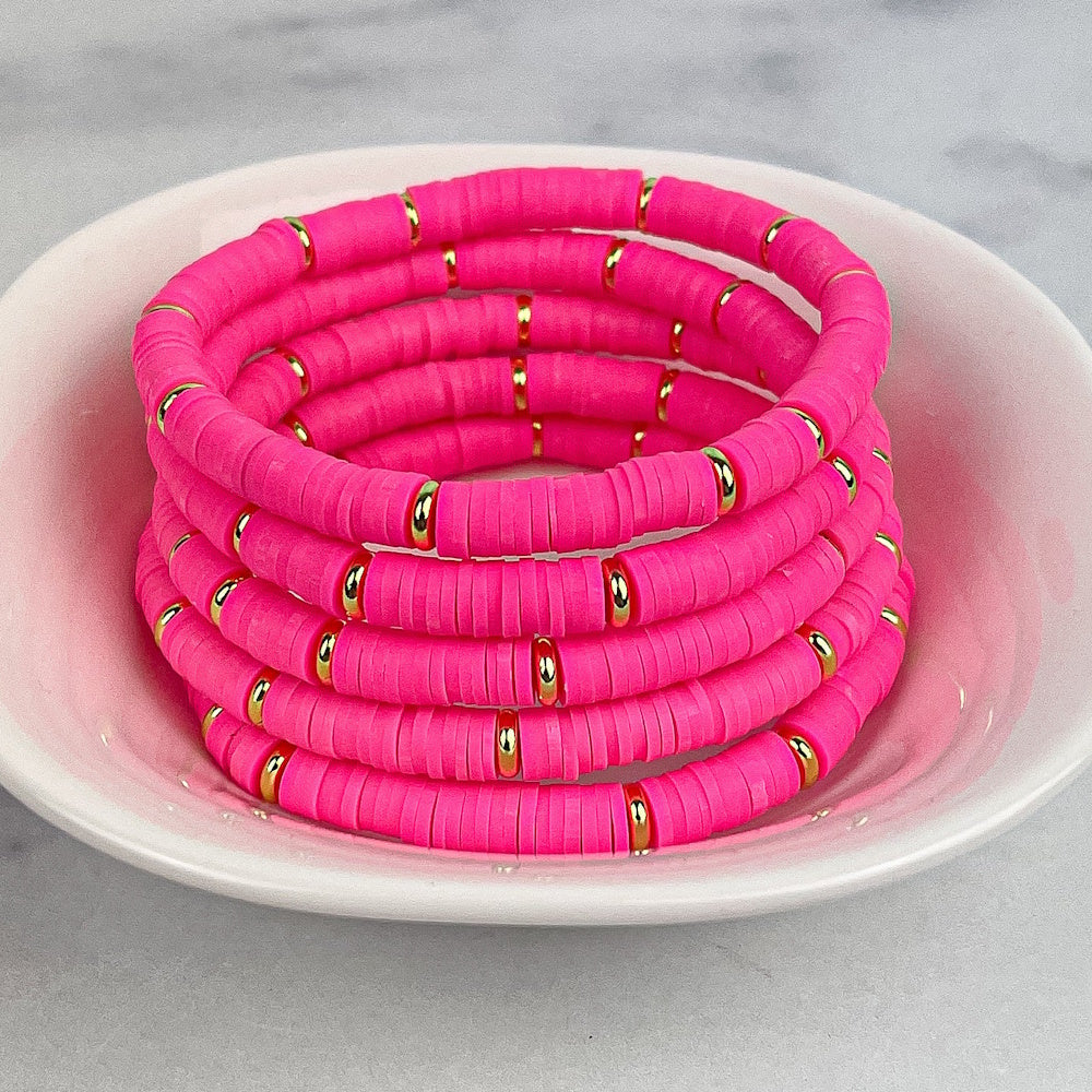 Stack of vibrant hot pink heishi bead bracelets with gold accents inside a small light pink dish.