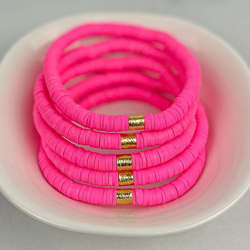 Stack of hot pink heishi bead bracelets, each with a single gold accent bead, in a white bowl.