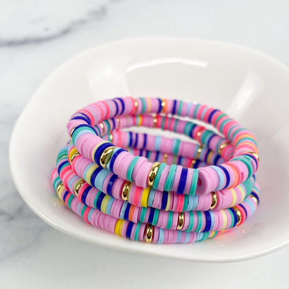 A stack of colorful heishi bead bracelets with lavender, rainbow, and gold accents in a white bowl.