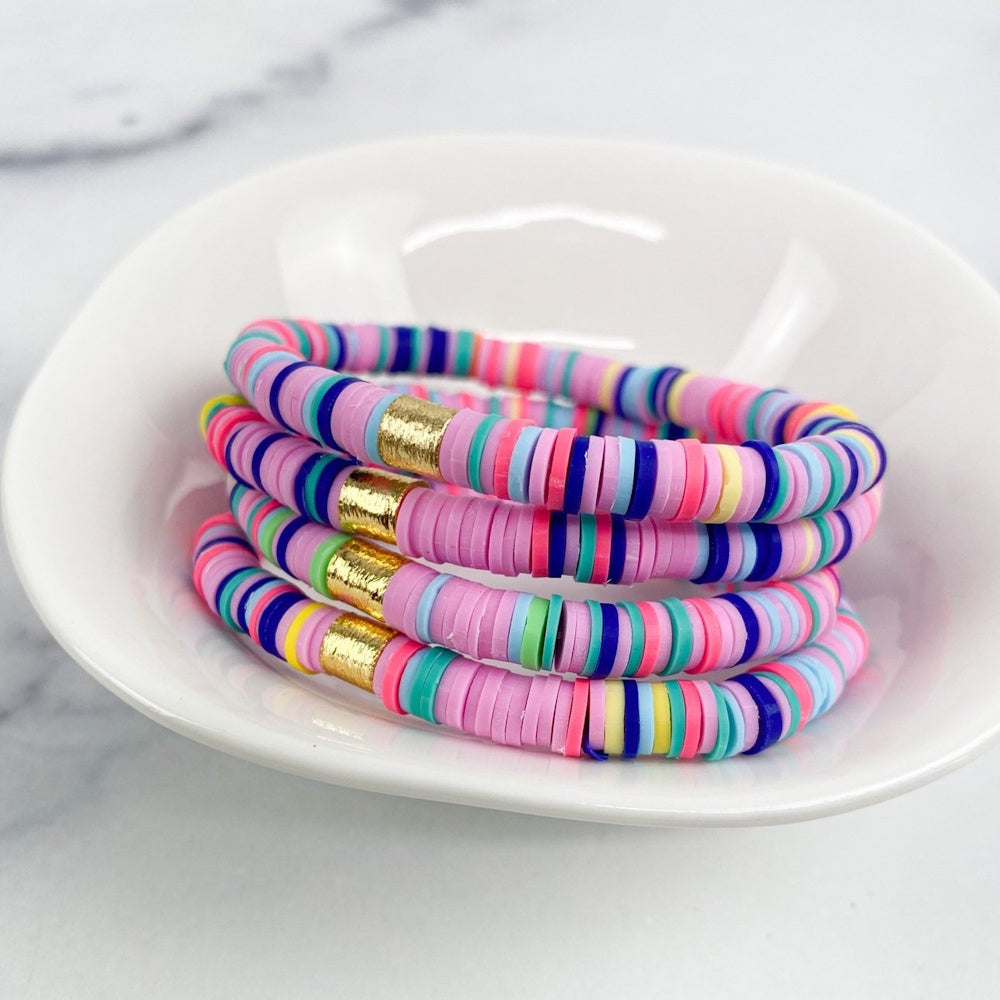 Stack of colorful rainbow heishi bead bracelets with gold accents inside a small white ceramic bowl.