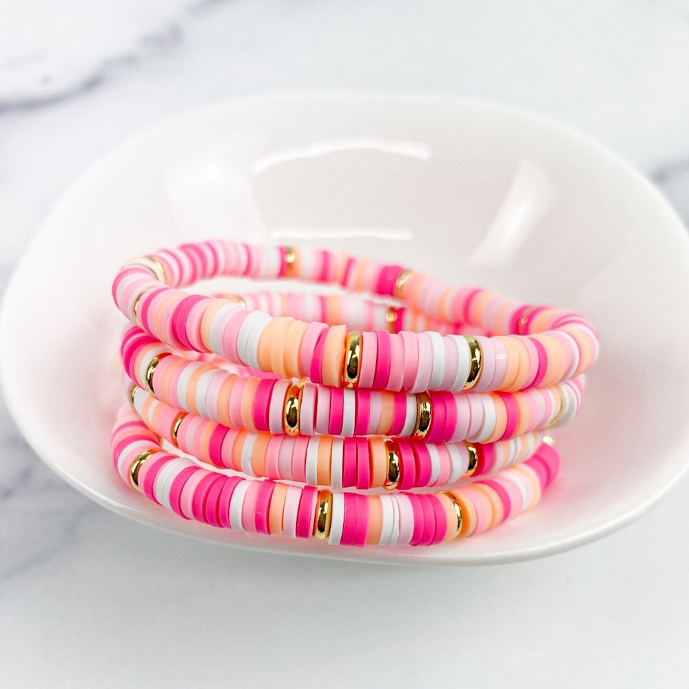 Stacked heishi bead bracelets in pink, peach, white, and gold accents inside a small white bowl.