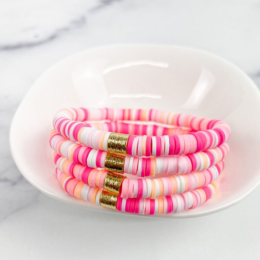 Four stacked pink, peach, and white heishi bead bracelets with gold accents resting in a small white bowl.