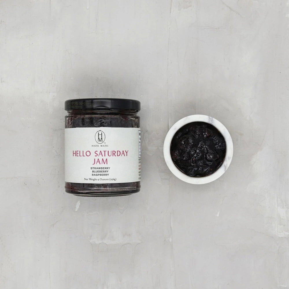 A jar of Hazel Mazel Hello Saturday Jam containing strawberry, blueberry, and raspberry next to a small bowl of jam.