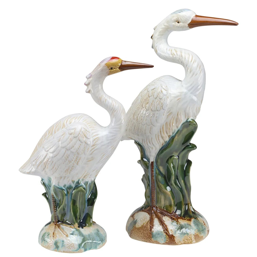 A pair of white heron figurines standing in green marsh grass, crafted with a glossy ceramic finish.