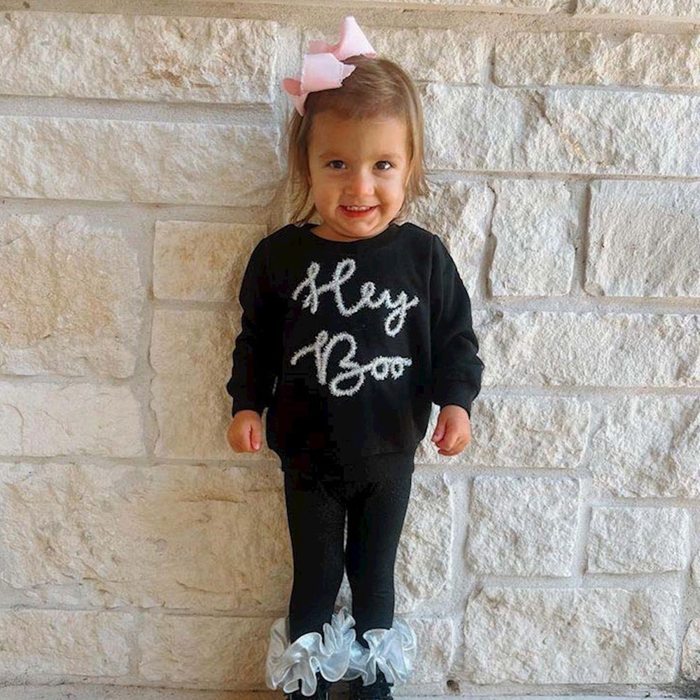 Smiling toddler girl in a black "Hey Boo" sweatshirt and leggings standing in front of a stone wall.