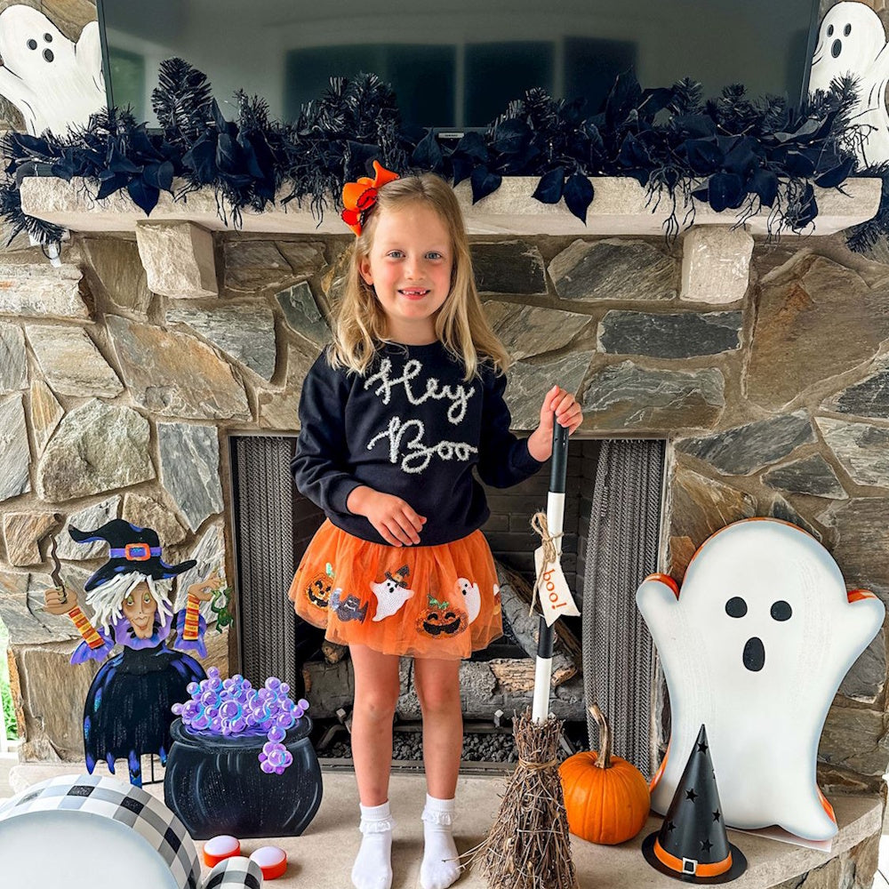 A young girl in a "Hey Boo" sweatshirt and orange tutu holds a broom in front of a decorated stone fireplace.