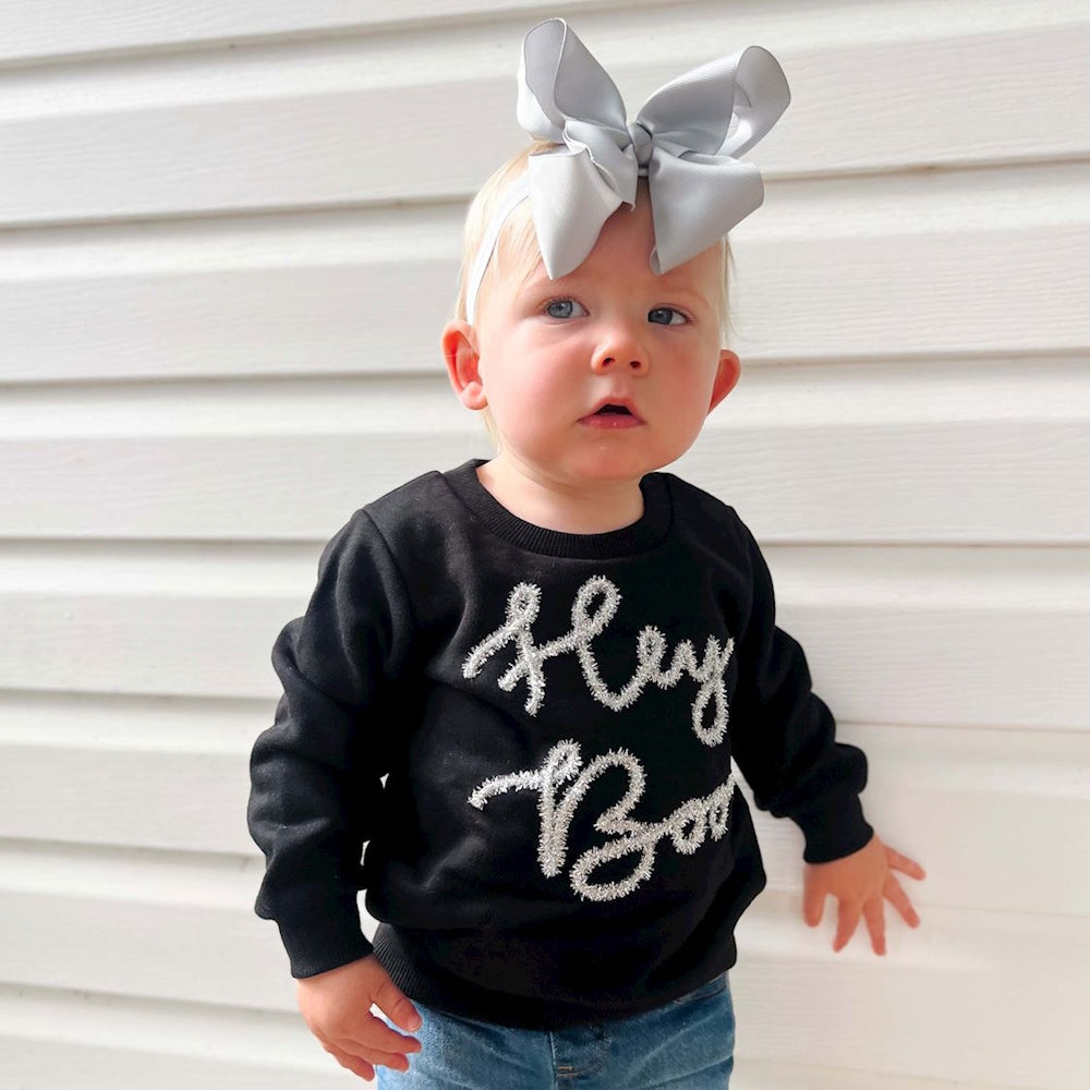 A toddler wearing a grey hairbow and a black "Hey Boo" sweatshirt stands against white siding.