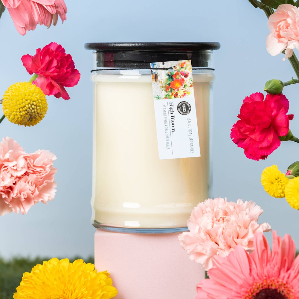 High Bloom jar candle by Bridgewater Candle Company surrounded by colorful pink and yellow flowers on a blue background.