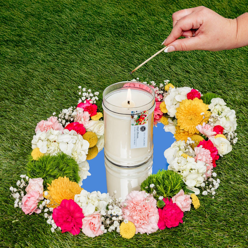 High Bloom scented jar candle being lit with a match, set in a vibrant floral wreath on grass.
