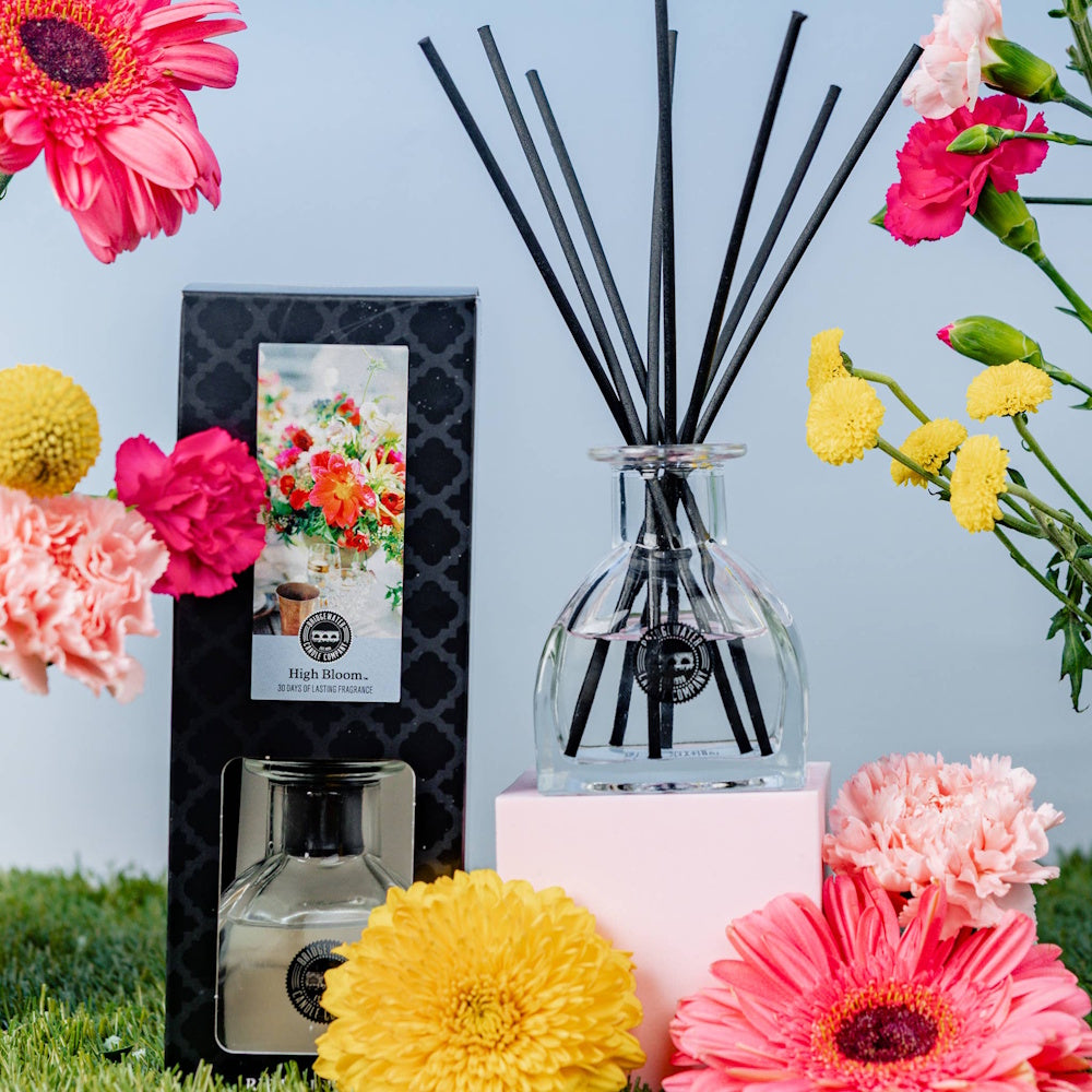 High Bloom reed diffuser and box set surrounded by pink and yellow flowers on grass with a blue background.