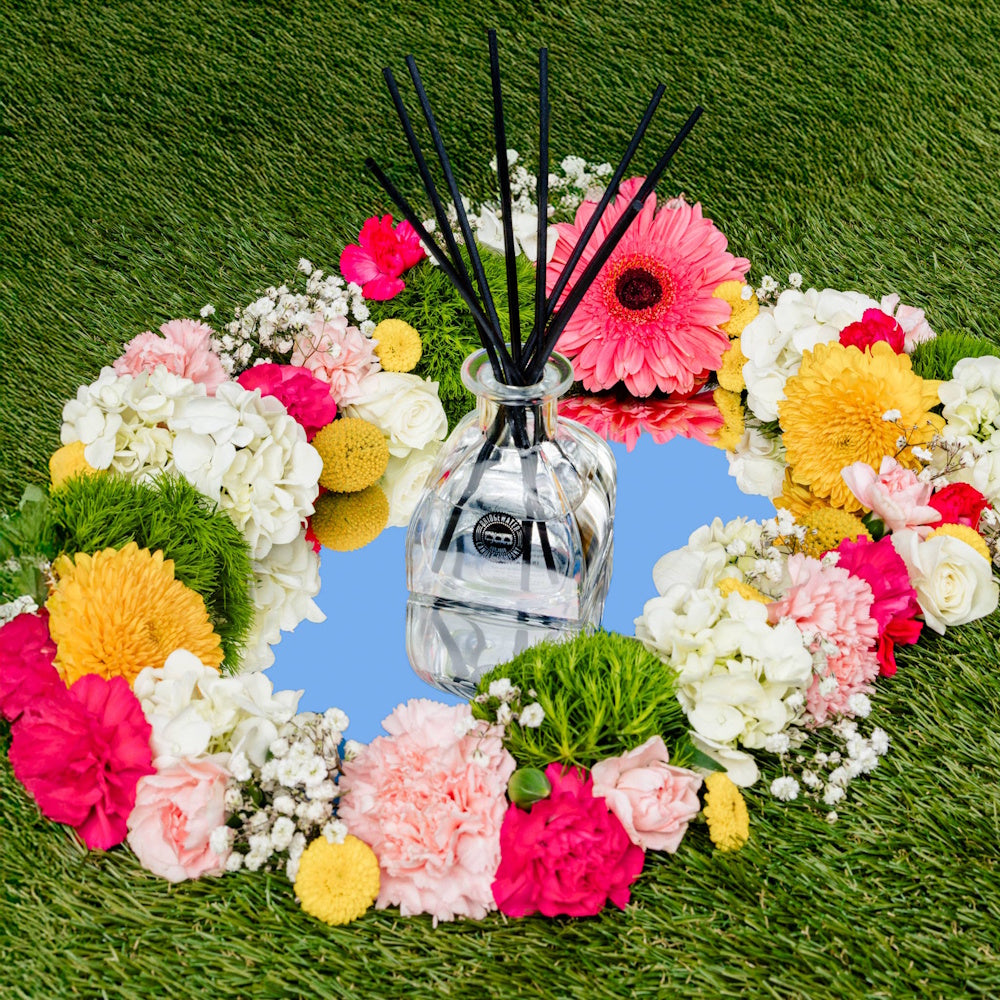 Glass reed diffuser on a mirror, surrounded by a colorful floral wreath of daisies and carnations on green grass.