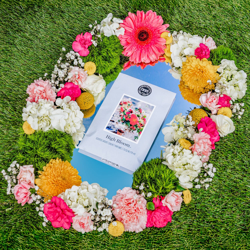 High Bloom scented sachet by Bridgewater Candle Company on a mirror surrounded by colorful spring flowers on green grass.