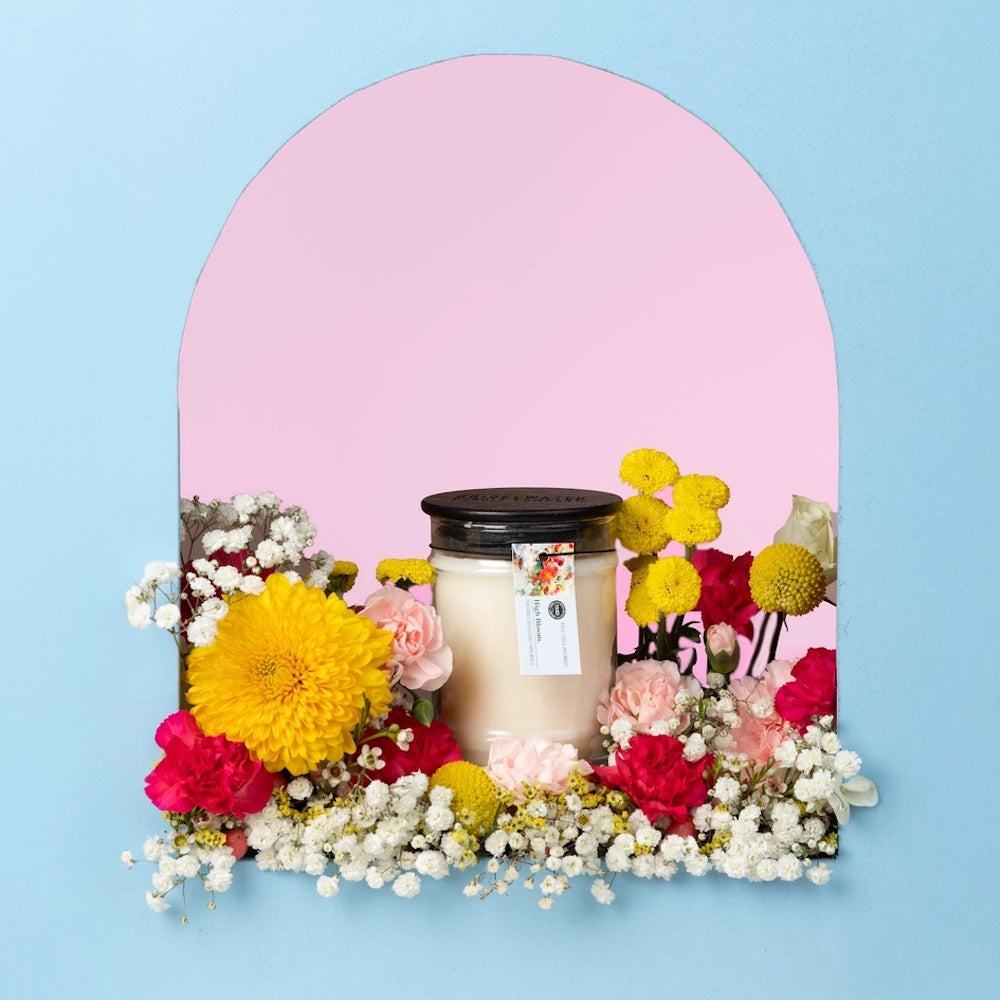 High Bloom candle framed by yellow chrysanthemums, pink carnations, and white baby's breath against a blue and pink backdrop.