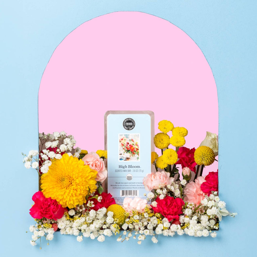 High Bloom scented wax bar surrounded by vibrant yellow, pink, and white spring flowers against a pink and blue background.