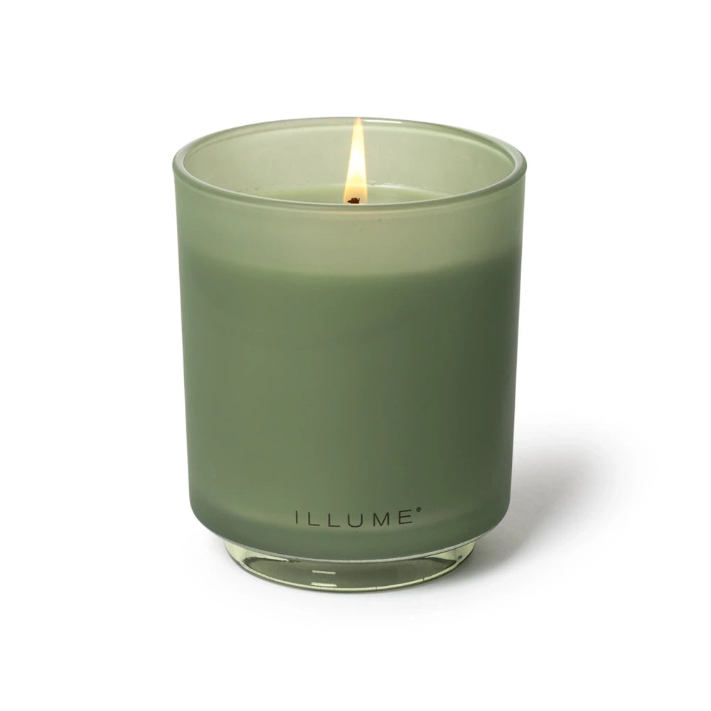 Lit Illume sage green candle glowing inside a frosted glass jar, set against a plain white background.