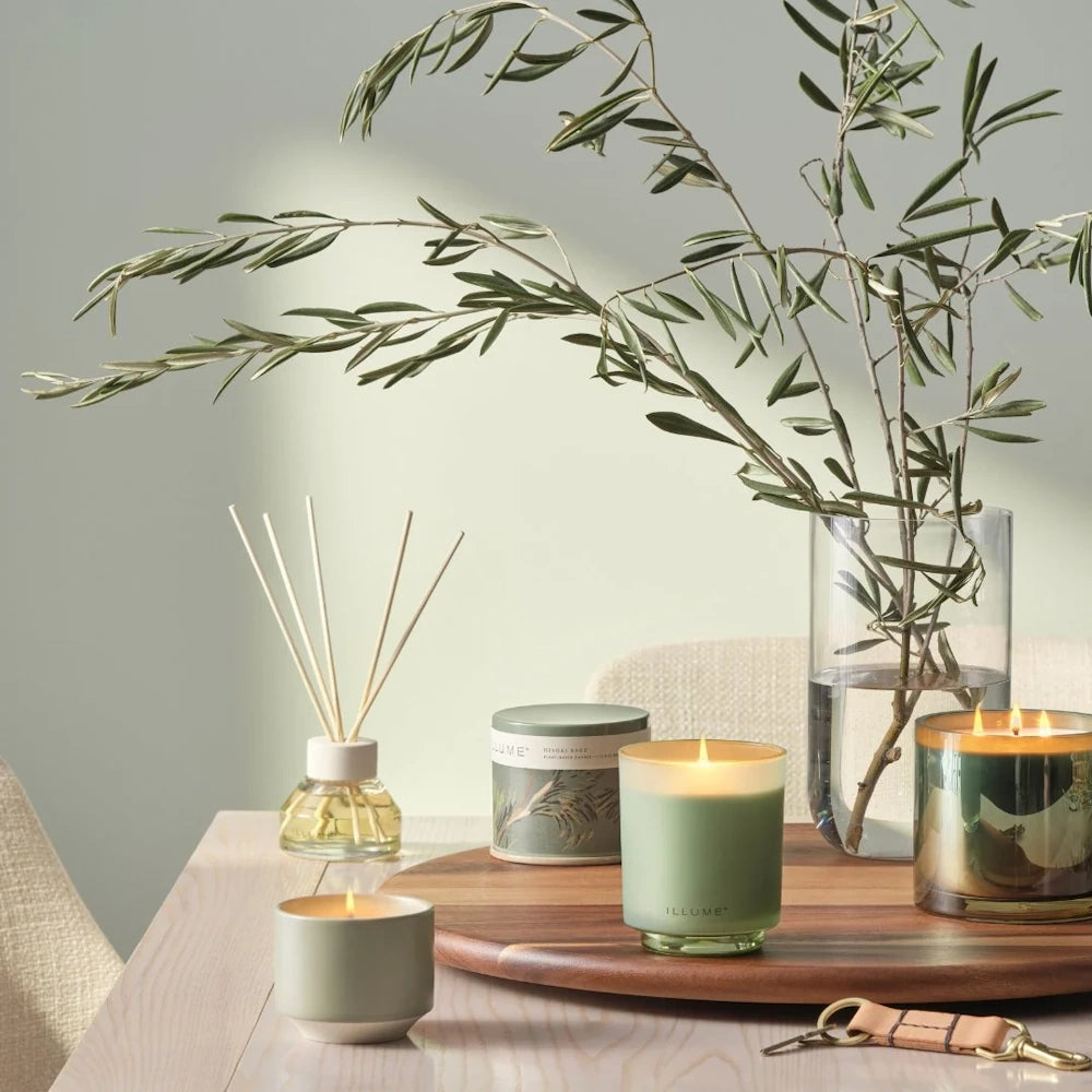 Illume Hinoki Sage candles, reed diffuser, and olive branches styled on a wooden tray atop a light table.