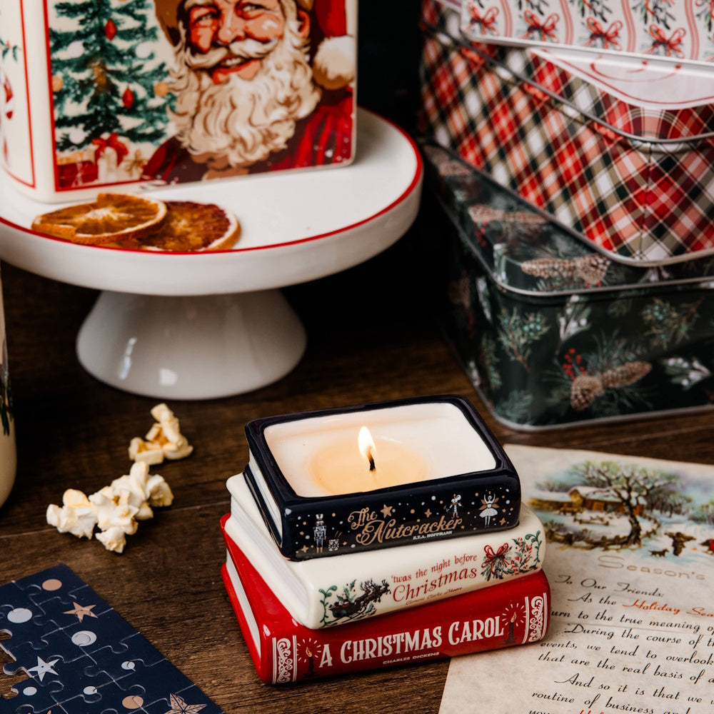 Lit candle shaped like a stack of Christmas book classics surrounded by holiday decor and festive tins.