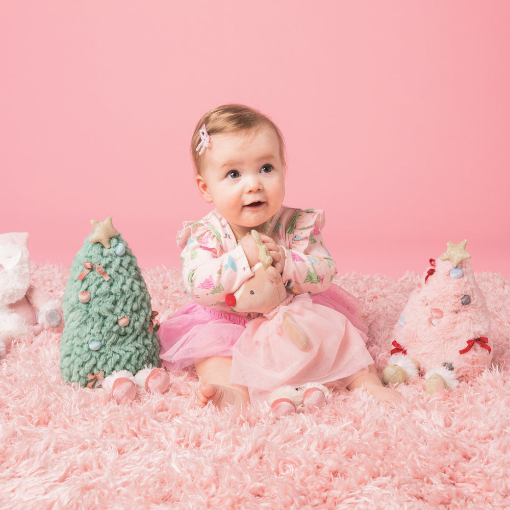 Baby in pink tutu holds plush reindeer, sitting on shag rug with decorative green and pink plush Christmas trees.