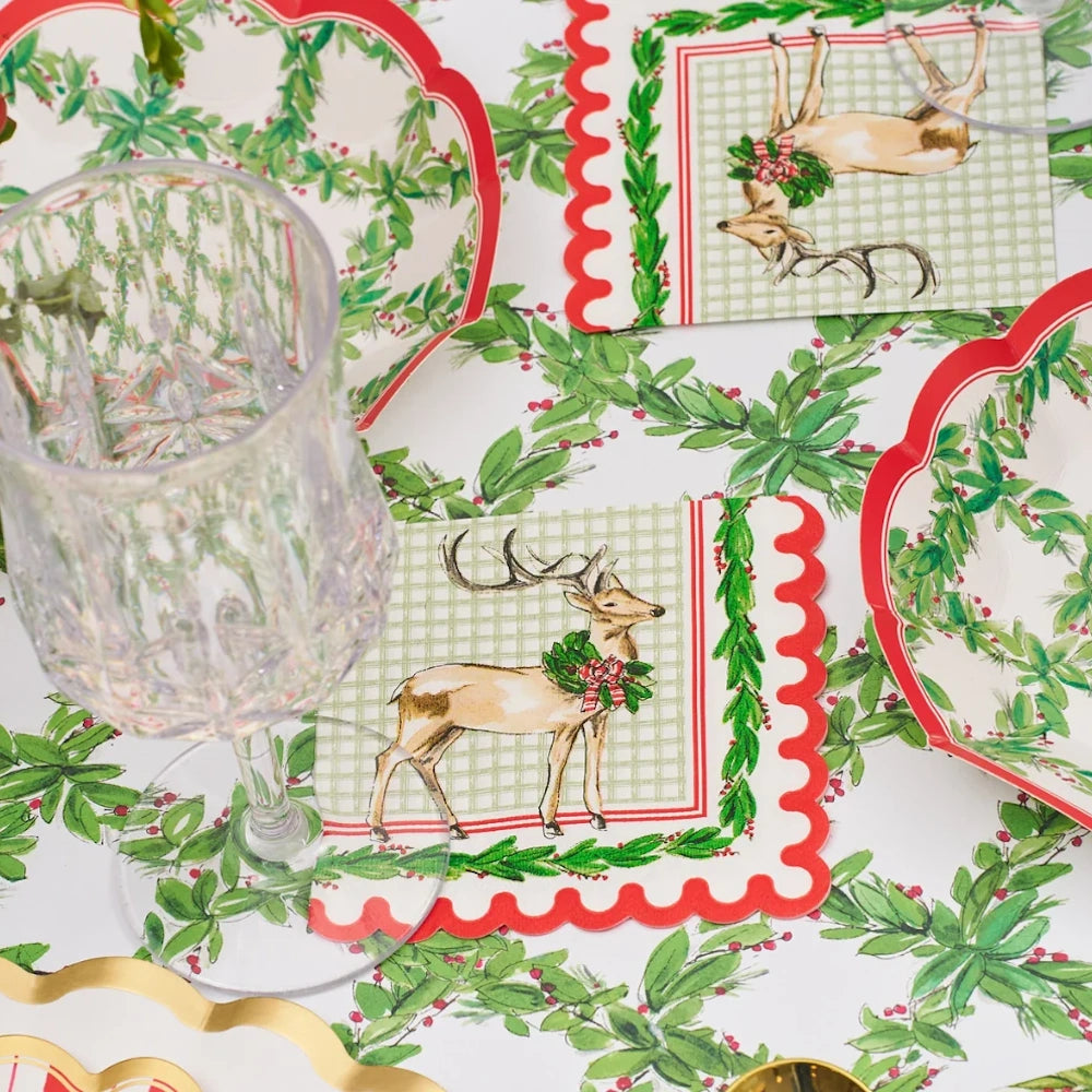 Lifestyle flat lay of a white paper cocktail napkin with a holiday deer, green plaid, and red border.