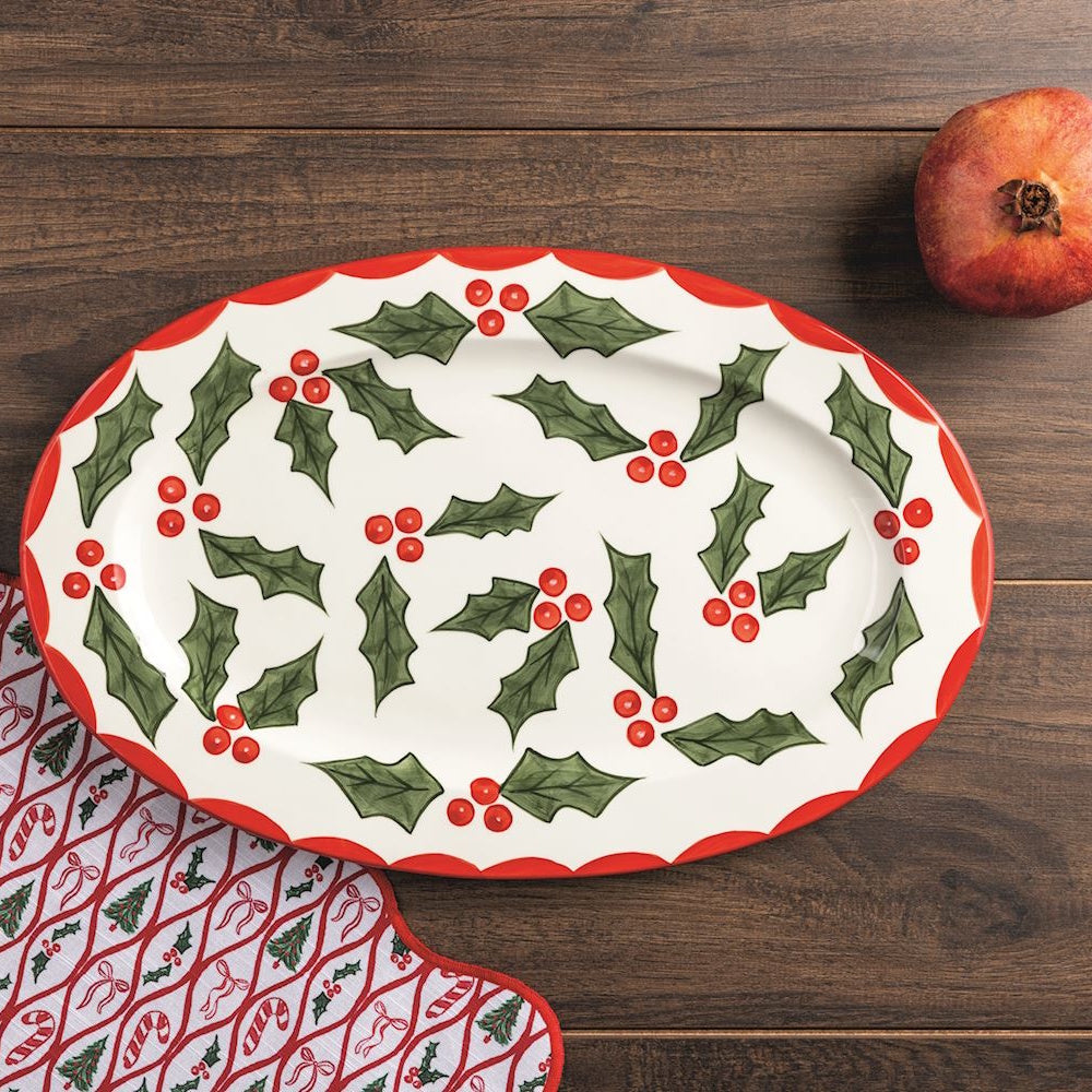 Oval holly leaf ceramic platter on a dark wood surface alongside a pomegranate and festive napkin.