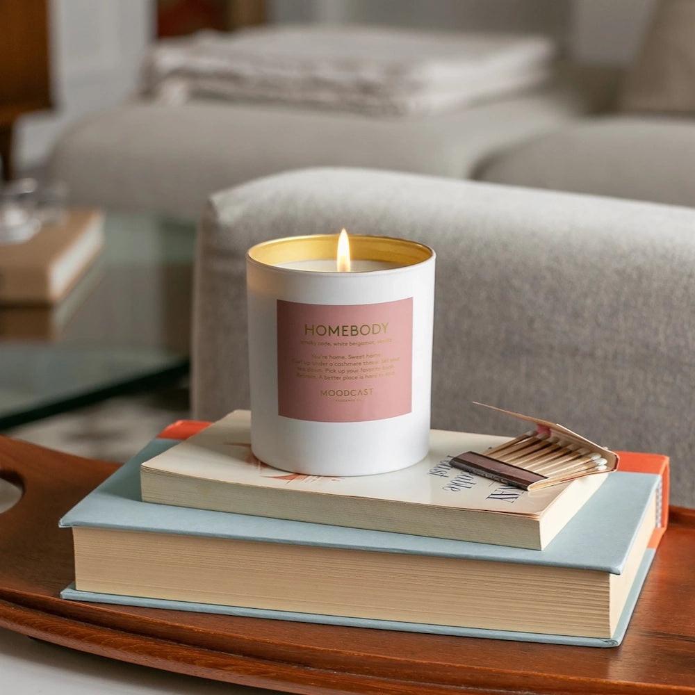 Lit Moodcast Homebody candle resting on a stack of books alongside an open matchbook.