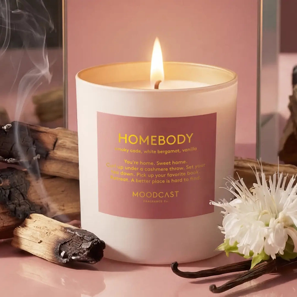 Lit white "Homebody" candle surrounded by charred wood, wisps of smoke, a white flower, and vanilla beans.