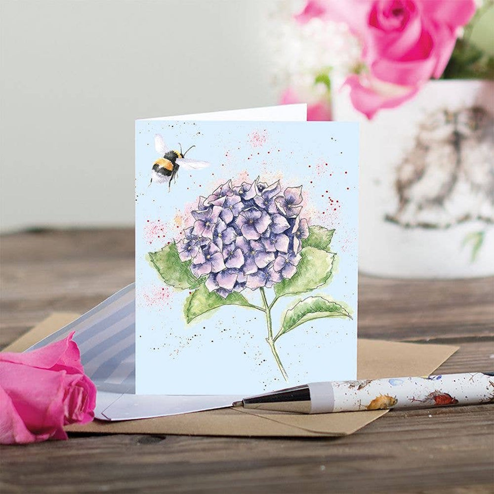 Watercolor illustration of a purple hydrangea and bumblebee on a blue gift card with a kraft envelope and pen.