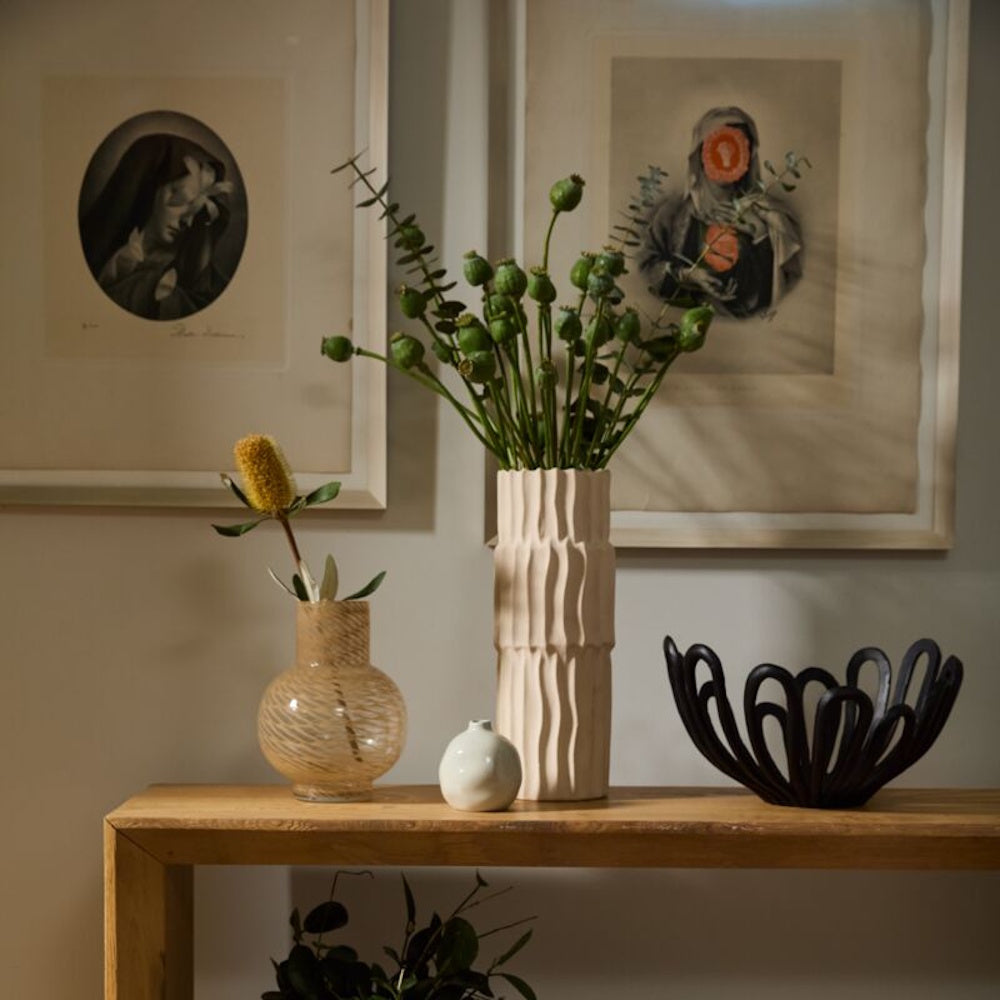 Minimalist home decor with textured ceramic vases, green botanicals, and a black wavy bowl on a wooden console table.
