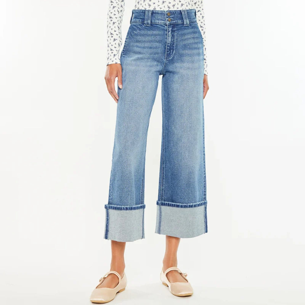 Model wears 90s-style cropped wide-leg blue jeans with large folded cuffs and beige flats against a white background.