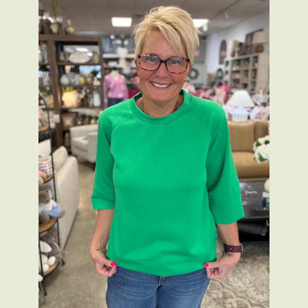 Woman in glasses wearing an island green 3/4 sleeve drop shoulder top and blue jeans in a boutique setting.
