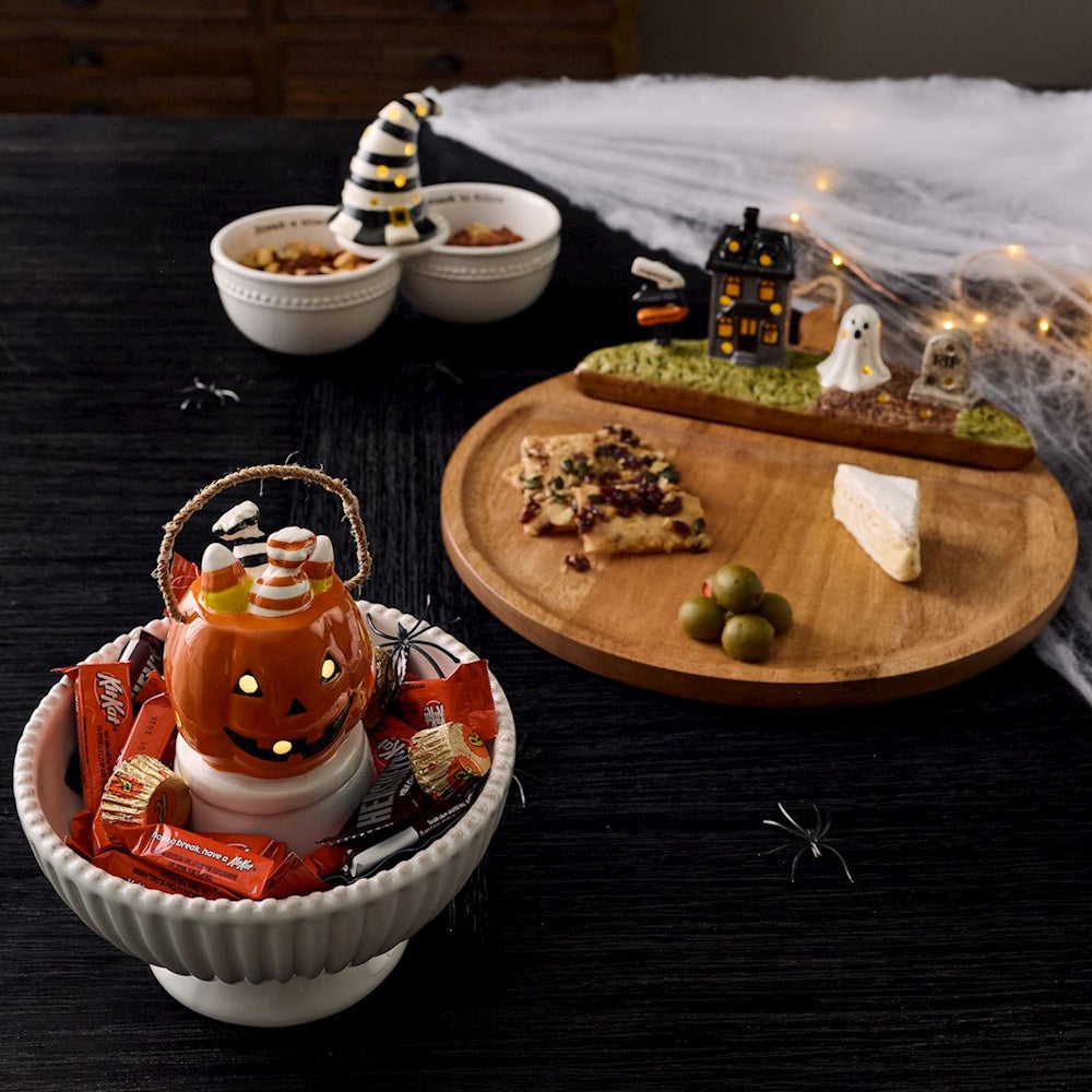 Lit Halloween jack-o'-lantern basket sitter on a white candy dish with Kit Kats and festive charcuterie board.