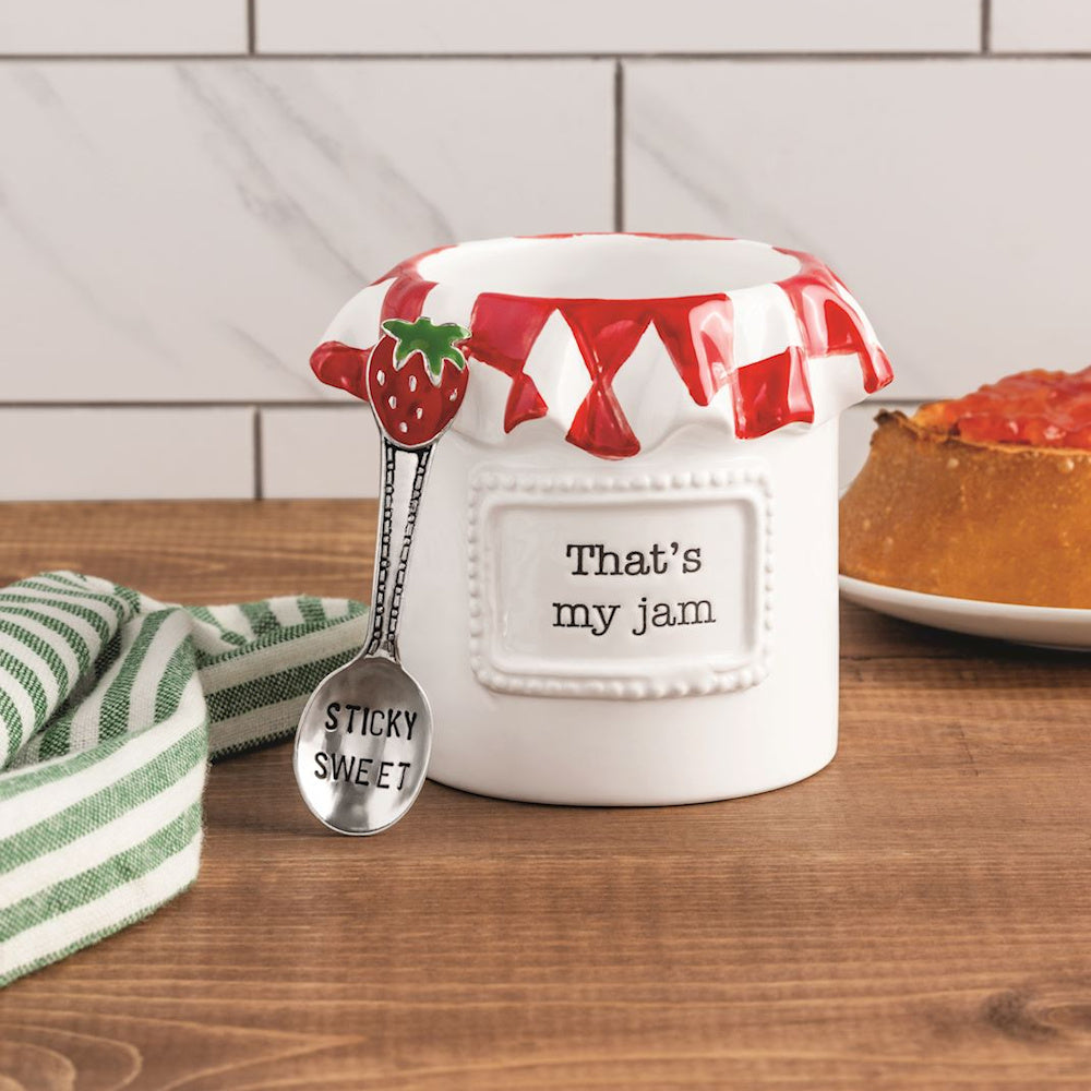 White ceramic "That's my jam" jar with a red dripped rim and a silver "Sticky Sweet" strawberry spoon on a wood surface.