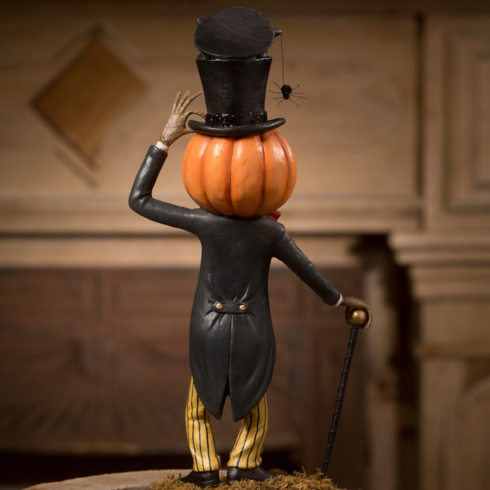 Back view of a pumpkin man figurine in a black tuxedo coat tipping his top hat, holding a cane.