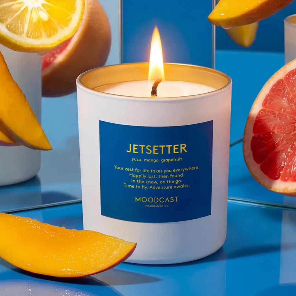 Lit Moodcast Jetsetter candle surrounded by sliced mangoes, grapefruit, and citrus against a blue backdrop.
