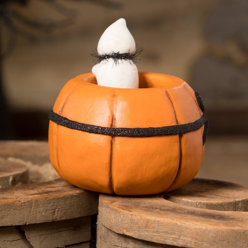 Back view of a small white ghost figure peeking out from an orange pumpkin decorated with black trim.