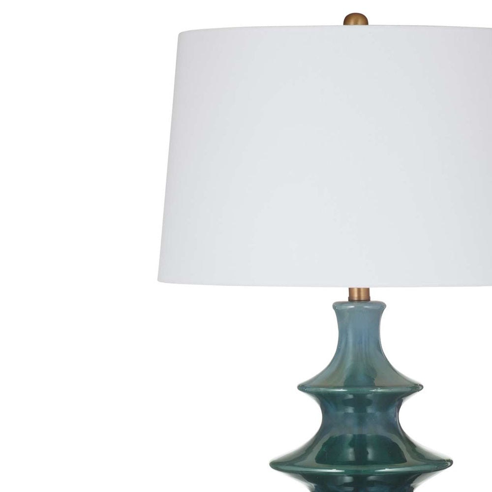 Close-up of a white drum lampshade with a brass finial atop a sculpted teal glass base.
