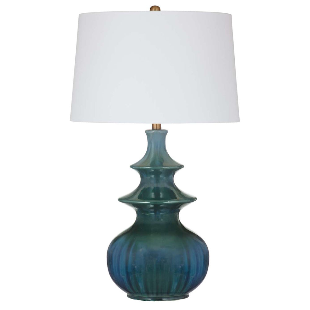 Table lamp with a tiered teal glass base and a white drum shade against a white background.