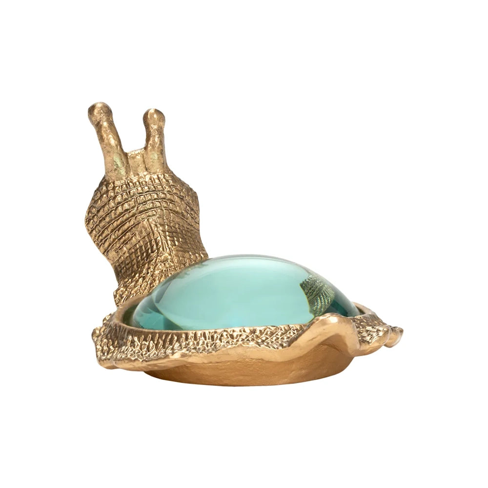 Back view of a brass snail figurine with a round glass lens set into its textured shell on a white background.