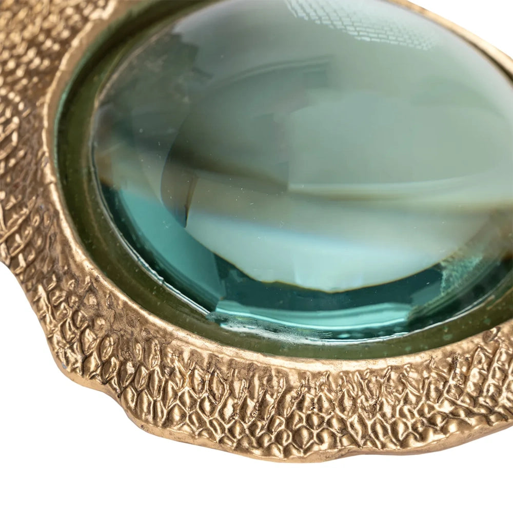 Close-up of a magnifying glass with clear glass set in a textured, antique gold frame with a scalelike pattern.