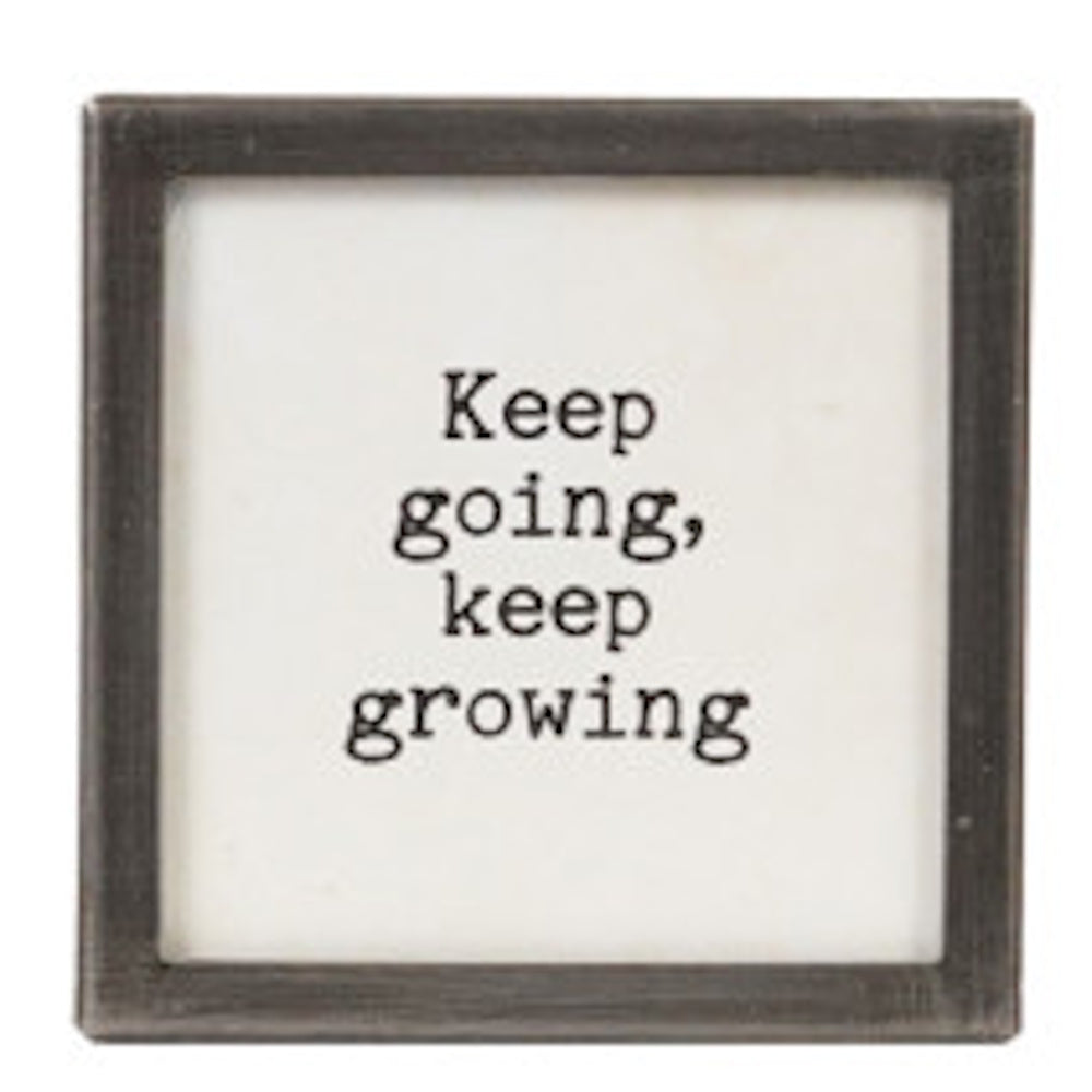 A square silver-framed sign with the text "Keep going, keep growing" in black typewriter font.