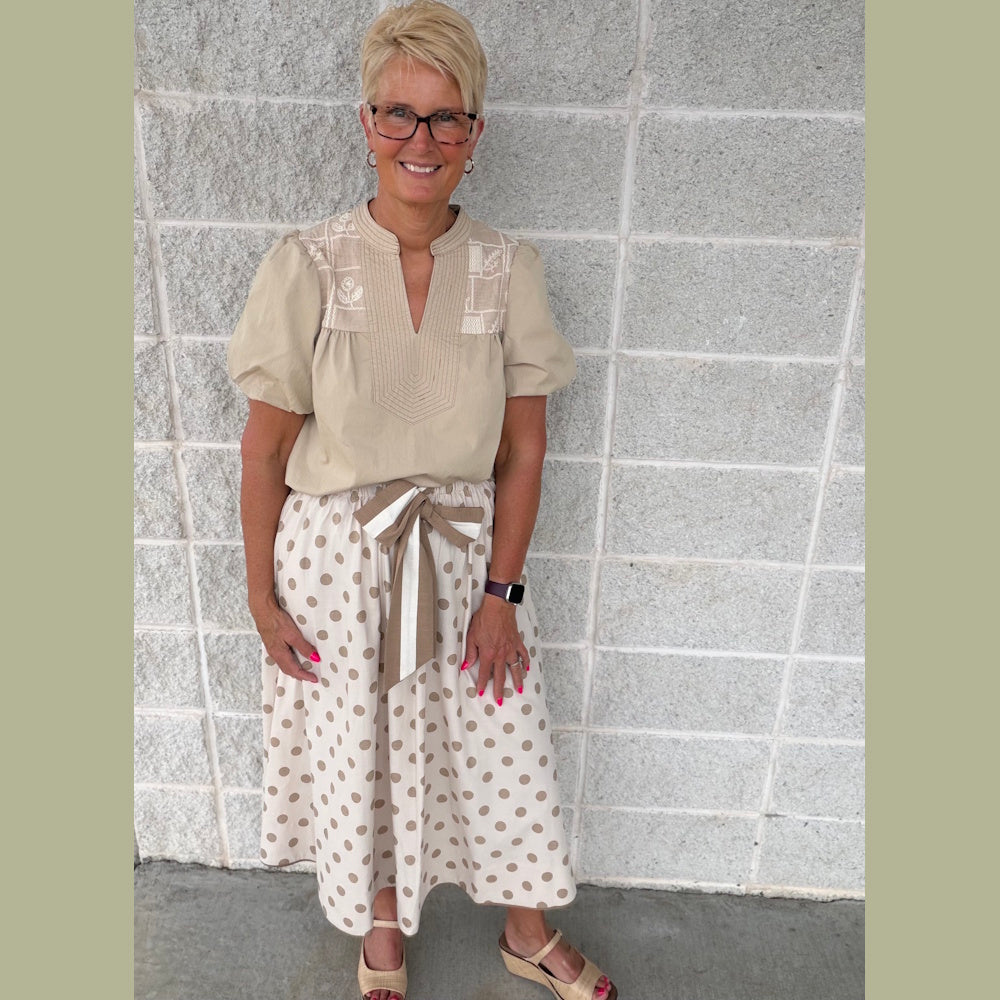 A woman with short blonde hair and glasses poses in a khaki puff-sleeve top and a polka-dot midi skirt.