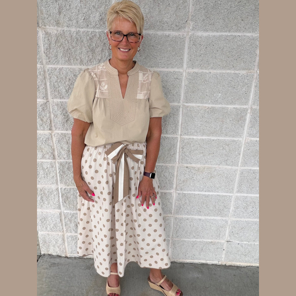 Smiling woman in a khaki puff-sleeve top and polka dot midi skirt with a bow belt posing against a gray stone wall.