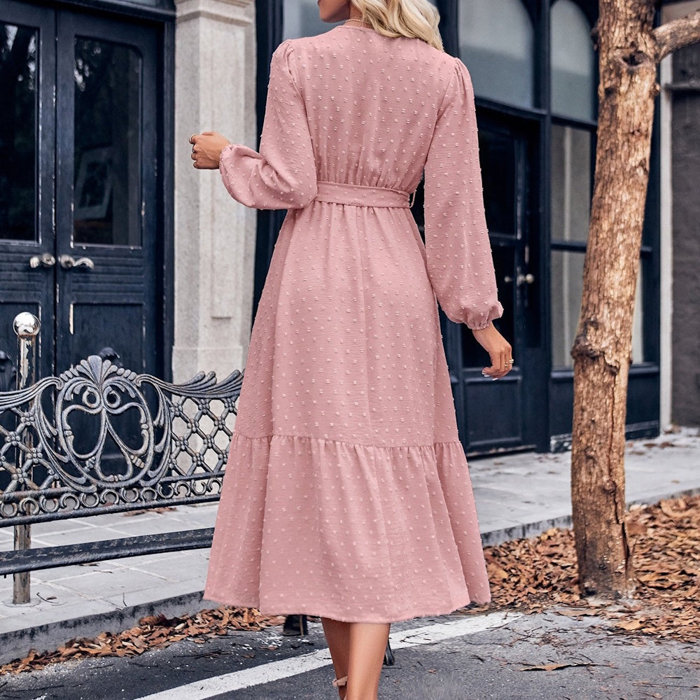 Rear view of a woman wearing a long-sleeved, dusty pink Swiss dot midi dress with a belted waist.