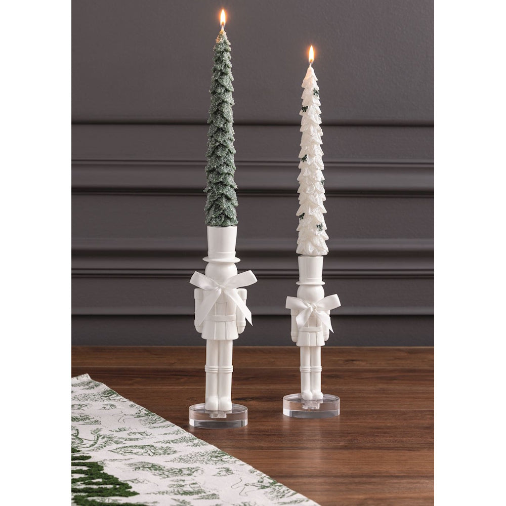 White nutcracker candlestick holders with bows holding lit tree-shaped taper candles on a wooden table.