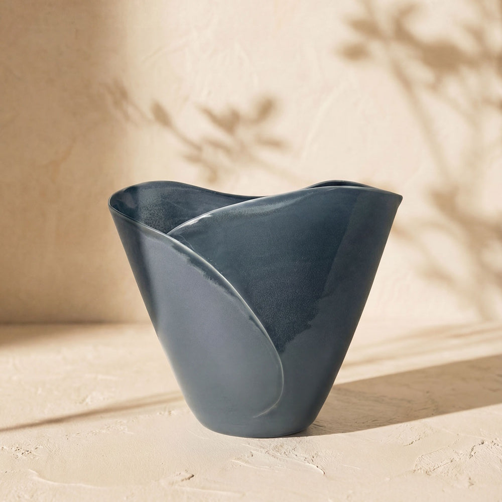 A slate blue ceramic bowl with a wavy, petal-like rim displayed against a neutral, sunlit wall with leafy shadows.
