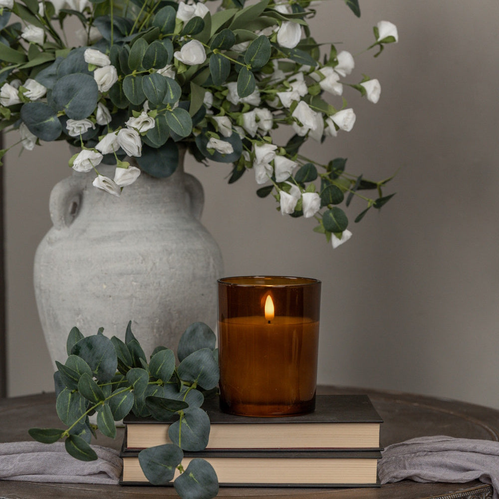 Amber glass candle with a lit flame atop stacked books next to eucalyptus greenery and a stone vase.