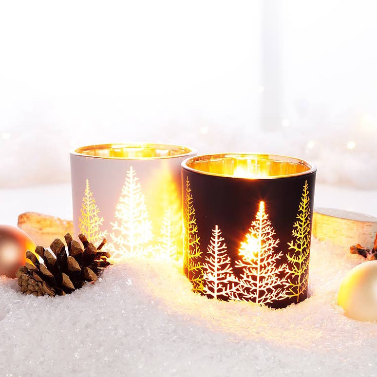 Glowing white and black glass votive candle holders with pine tree cutouts in a snowy scene.