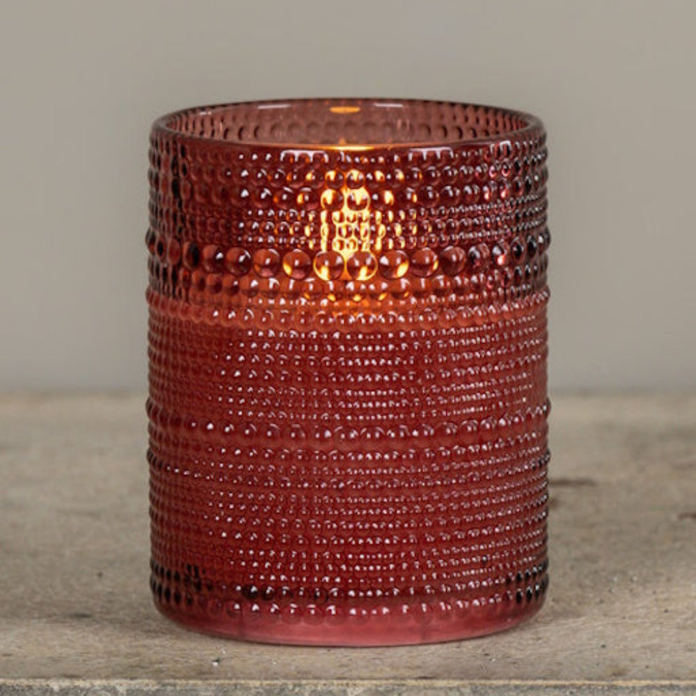 Lit candle inside a burgundy dotted glass vessel sitting on a stone surface against a neutral wall.