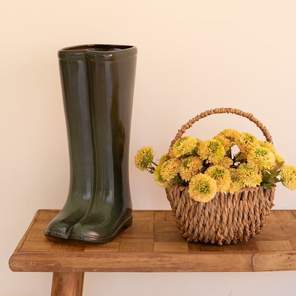 Large dark green ceramic boot vase next to a woven flower basket on a wooden bench.