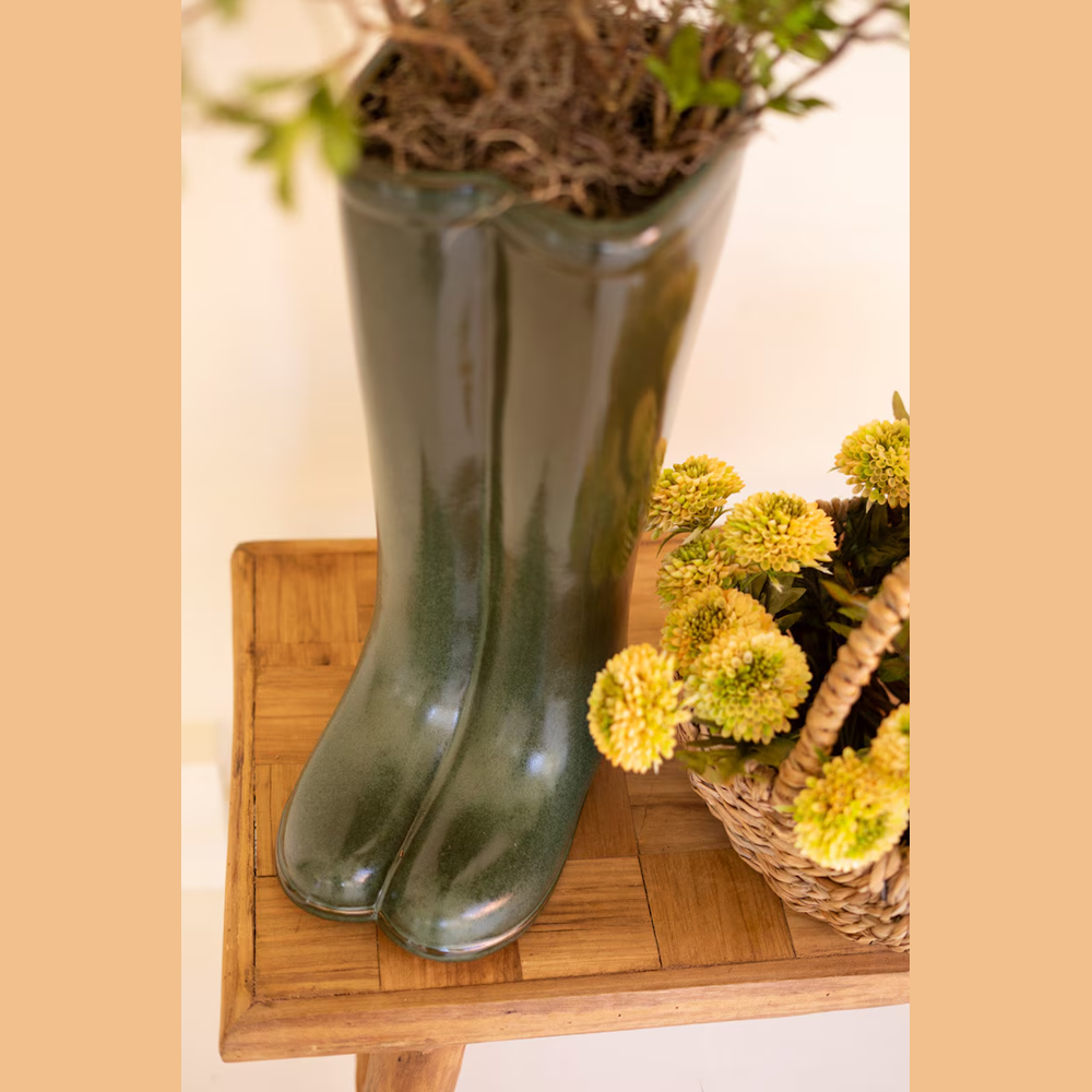 Large dark green ceramic rain boot vase with plants on a wooden table next to a basket of yellow flowers.